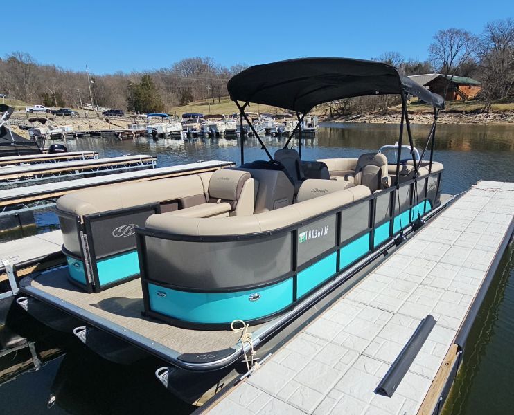 Turquoise-and-gray pontoon boat with black canopy moored at a lakeside marina dock, beige cushioned seating and calm water under a clear blue sky.