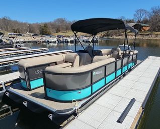 Turquoise-and-gray pontoon boat with black canopy moored at a lakeside marina dock, beige cushioned seating and calm water under a clear blue sky.