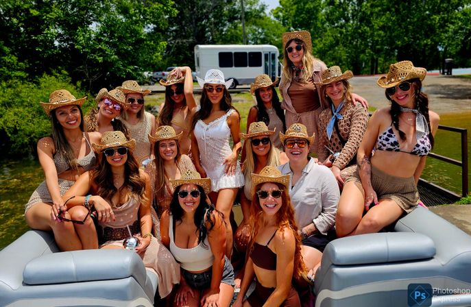 Happy group of women wearing straw cowboy hats and sunglasses posing on a pontoon boat at a sunny lakeside dock with trees in the background