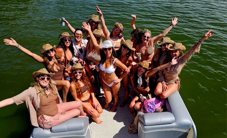 Group of women in swimsuits and cowboy hats partying on a pontoon boat over green lake water, arms raised and smiling with drinks — sunny lake boat party vibe