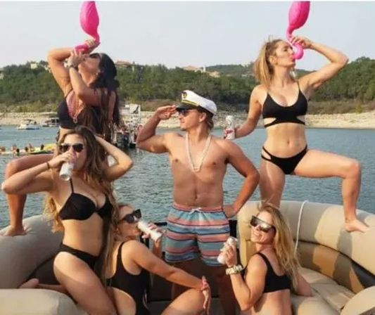 Group of six adults on a pontoon boat at a sunny lake: shirtless man in a captain's hat poses while five women in black bikinis sip canned drinks and use pink flamingo drink holders, tree-lined shore in the background.