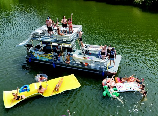 Aerial view of a summer lake party with a pontoon boat, people cheering on deck, colorful inflatables including a large yellow floating mat and pool loungers on green water near a tree-lined shore.