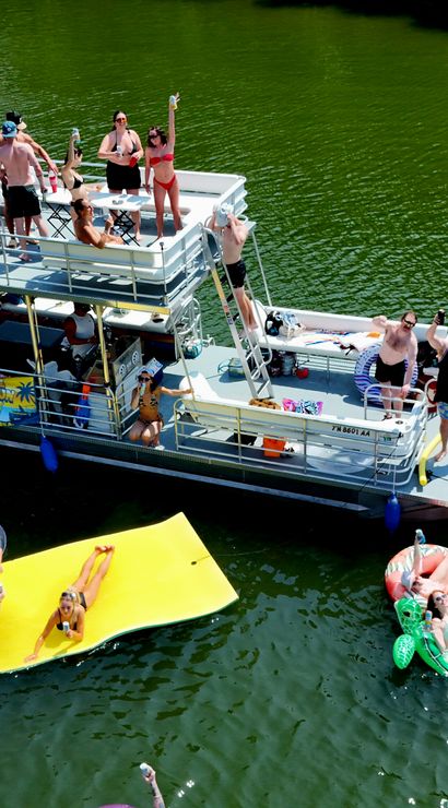 Aerial view of a summer lake party with a pontoon boat, people cheering on deck, colorful inflatables including a large yellow floating mat and pool loungers on green water near a tree-lined shore.