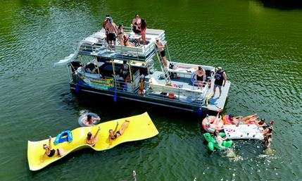 Aerial shot of a pontoon boat on a green lake during summer, with people sunbathing on the deck and relaxing on a large yellow floating mat and colorful inflatables nearby.