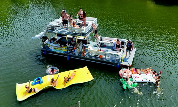 Aerial shot of a pontoon boat on a green lake during summer, with people sunbathing on the deck and relaxing on a large yellow floating mat and colorful inflatables nearby.
