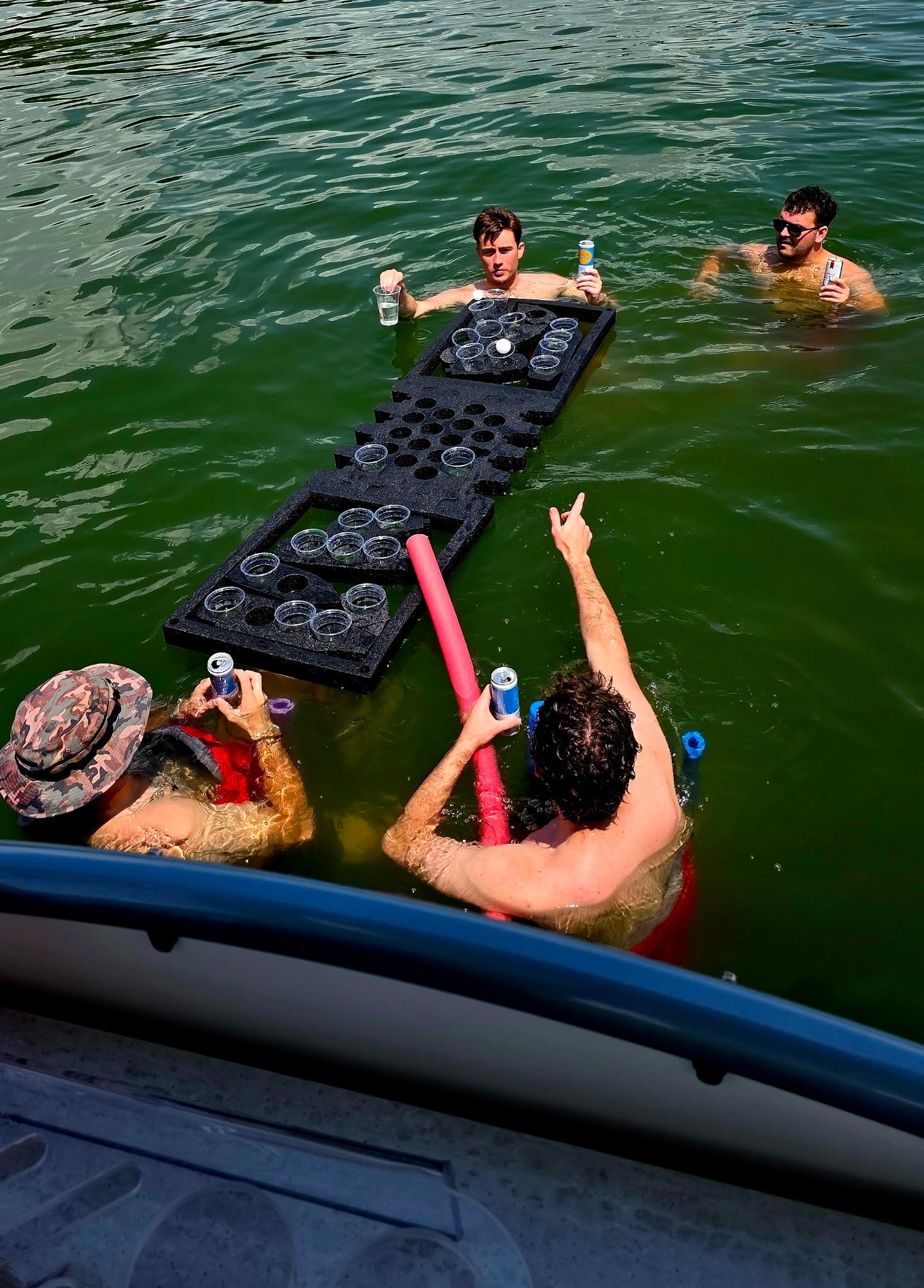 Four friends in a green lake playing floating beer‑pong on a foam cup board beside a boat, holding canned drinks and a pool noodle