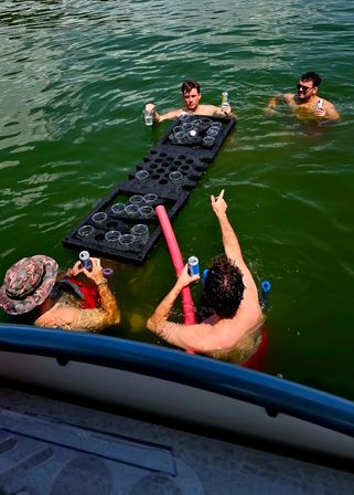 Four friends in a green lake playing floating beer‑pong on a foam cup board beside a boat, holding canned drinks and a pool noodle