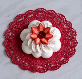 White flower-shaped meringue pavlova topped with piped cream and sliced strawberries, served on a red lace doily on a white marble countertop
