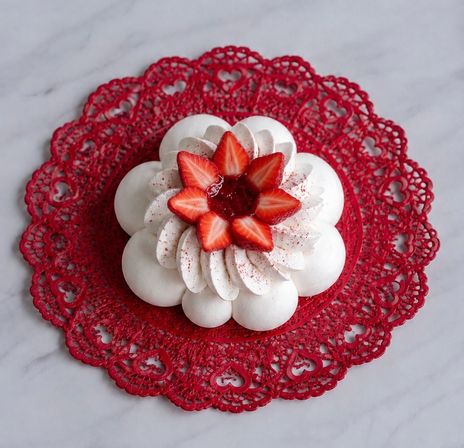 White flower-shaped meringue pavlova topped with piped cream and sliced strawberries, served on a red lace doily on a white marble countertop