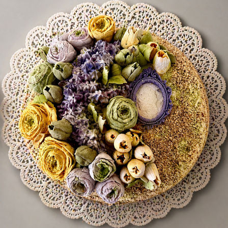 Top-down view of a round buttercream floral cake decorated with pastel piped roses, buds and textured frosting, sitting on a lace paper doily — artisan dessert centerpiece
