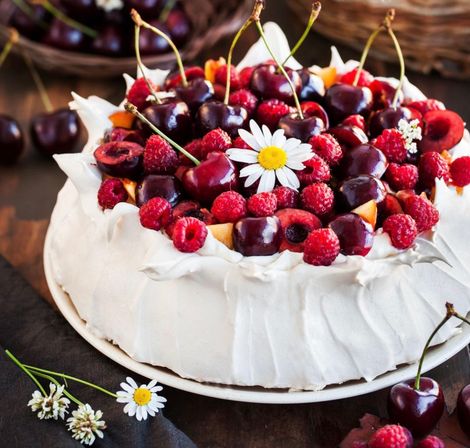 Fluffy white pavlova topped with glossy dark cherries, bright raspberries and a daisy, styled on a rustic wooden table