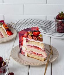 Tempting slice of layered strawberry cream cake topped with clear berry jelly, fresh strawberries, cherries and edible flowers on a beige plate with a gold fork on a white wooden table.