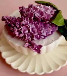 Close-up of purple lilac blossoms crowning a small white frosted cake on a scalloped white plate against a soft pink background.
