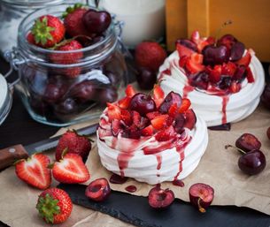 Two mini pavlovas on parchment-lined slate topped with chopped strawberries, halved cherries and glossy berry sauce with a jar of fresh berries nearby — bright summer dessert.