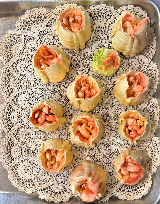 Metal baking tray lined with vintage lace doilies holding a dozen peach-and-cream piped buttercream flower buds, with one pale green bud, arranged in neat rows
