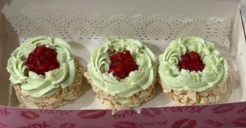 Three pistachio-green meringue nests piped with pale green cream and filled with raspberry jam, arranged in a pink bakery box on a paper doily