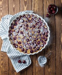 Homemade cherry clafoutis in a fluted pie dish dusted with powdered sugar, set on a striped kitchen towel over a rustic wooden table with a bowl of cherries and a flour sifter nearby