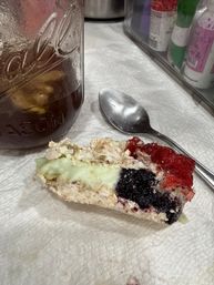 Close-up breakfast scene: mason jar of iced tea with ice, a metal spoon on a paper towel, and a slice of toast topped with green spread and blackberry and strawberry jam.