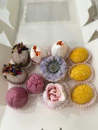 Box of colorful, floral-inspired handmade confections in paper liners — pink rose-shaped sweets, cracked white meringue “eggs,” purple flower cookie, chocolate truffle with dried petals and yellow coconut-coated bites