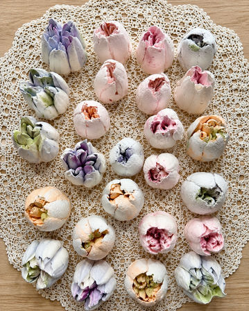 Pastel meringue cookies shaped like peony buds arranged on a crocheted lace doily over a wooden tabletop — colorful shabby‑chic dessert display
