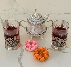 Two ruby-red tea glasses in ornate metal holders flanking a pearl-studded silver sugar bowl on a white marble countertop, with pink and orange flower-shaped cookies in front.