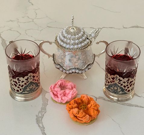Two ruby-red tea glasses in ornate metal holders flanking a pearl-studded silver sugar bowl on a white marble countertop, with pink and orange flower-shaped cookies in front.