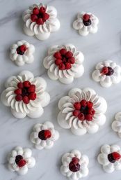 Mini pavlova-style meringue desserts piped into floral shapes and topped with raspberry jam and fresh raspberries, arranged on a white marble surface.
