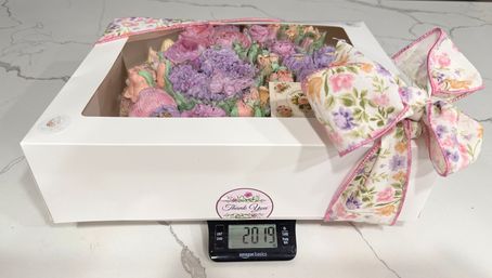 White bakery box with clear window revealing pastel buttercream rose cupcakes, tied with a floral ribbon bow, sitting on a digital kitchen scale showing 2.019 on a marble countertop