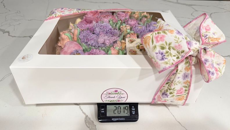 White bakery box with clear window revealing pastel buttercream rose cupcakes, tied with a floral ribbon bow, sitting on a digital kitchen scale showing 2.019 on a marble countertop