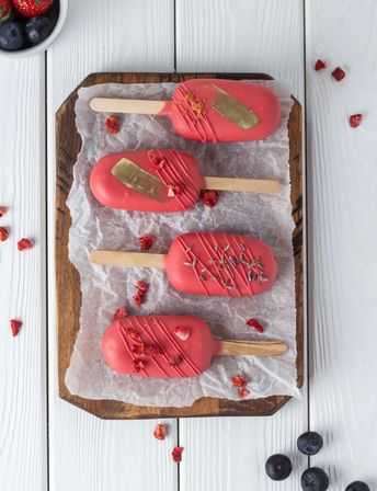 Four pink chocolate‑coated ice cream bars on a parchment‑lined wooden board, decorated with gold brushstrokes and dried berries, fresh blueberries scattered on a white wooden table.