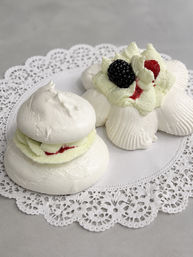 Close-up of two white meringue pavlova desserts on a lace paper doily, layered with whipped cream and raspberry jam, topped with a fresh blackberry and raspberry