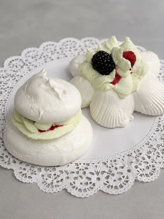 Close-up of two white meringue pavlova desserts on a lace paper doily, layered with whipped cream and raspberry jam, topped with a fresh blackberry and raspberry