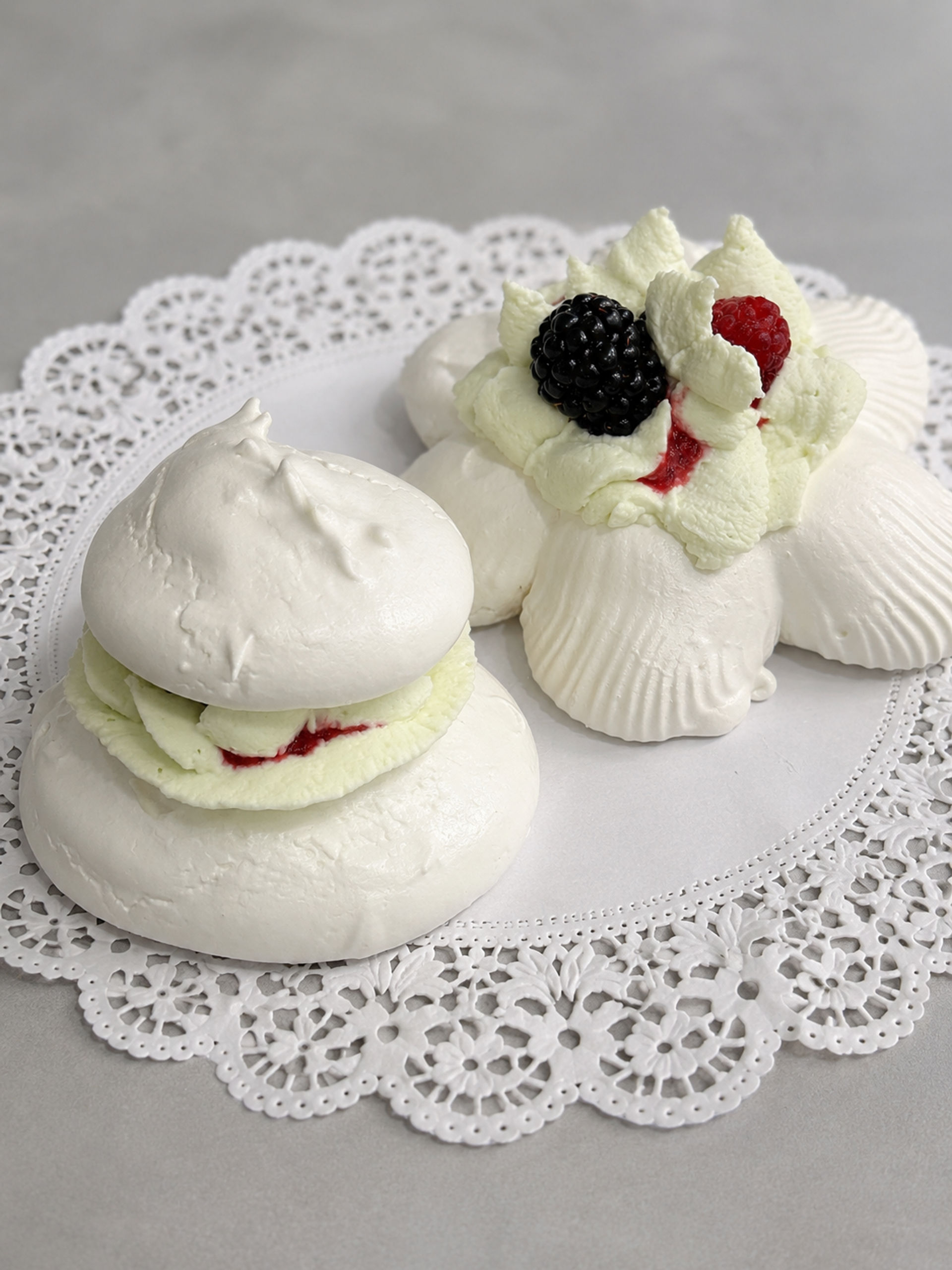 Close-up of two white meringue pavlova desserts on a lace paper doily, layered with whipped cream and raspberry jam, topped with a fresh blackberry and raspberry