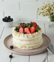 Layered pavlova dessert topped with halved strawberries, blueberries and fresh mint on a neutral plate on a white wooden table with chamomile flowers in the background