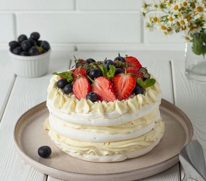 Layered pavlova dessert topped with halved strawberries, blueberries and fresh mint on a neutral plate on a white wooden table with chamomile flowers in the background