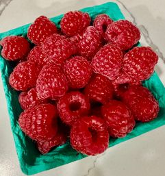 Close-up of juicy red raspberries piled in a teal berry carton on a light marble countertop.