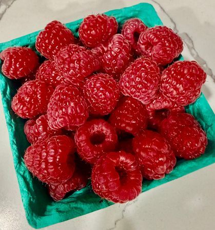 Close-up of juicy red raspberries piled in a teal berry carton on a light marble countertop.