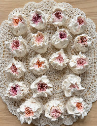 Close-up of peony-inspired pastel meringue cookies with ruffled white edges and pink-peach centers arranged on a vintage crochet doily over a light wooden table