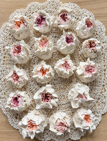 Close-up of peony-inspired pastel meringue cookies with ruffled white edges and pink-peach centers arranged on a vintage crochet doily over a light wooden table
