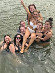Happy group of friends in swimsuits lifting a smiling woman in a white bikini while splashing in shallow lake water — lively summer outing