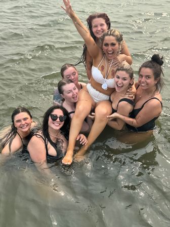 Happy group of friends in swimsuits lifting a smiling woman in a white bikini while splashing in shallow lake water — lively summer outing