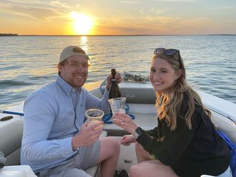 Smiling friends toasting with plastic cups and a wine bottle on a motorboat at golden sunset over calm lake waters