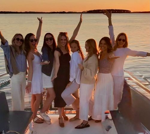 Group of eight women friends posing on a boat at golden sunset over calm coastal waters, arms raised and smiling in summer dresses and casual outfits.