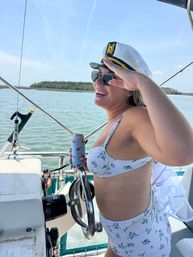 Bride-to-be in floral bikini and 'Bride' captain's hat saluting while steering a boat, holding a drink in a flamingo koozie with calm coastal waters and tree-lined shore in the background.