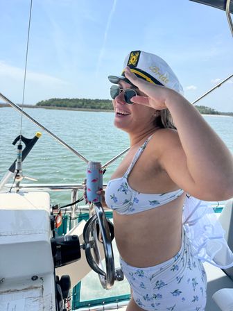 Bride-to-be in floral bikini and 'Bride' captain's hat saluting while steering a boat, holding a drink in a flamingo koozie with calm coastal waters and tree-lined shore in the background.