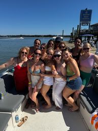 Group of friends posing on a boat at a sunny marina, wearing swimsuits and sunglasses with calm harbor water and docks in the background.