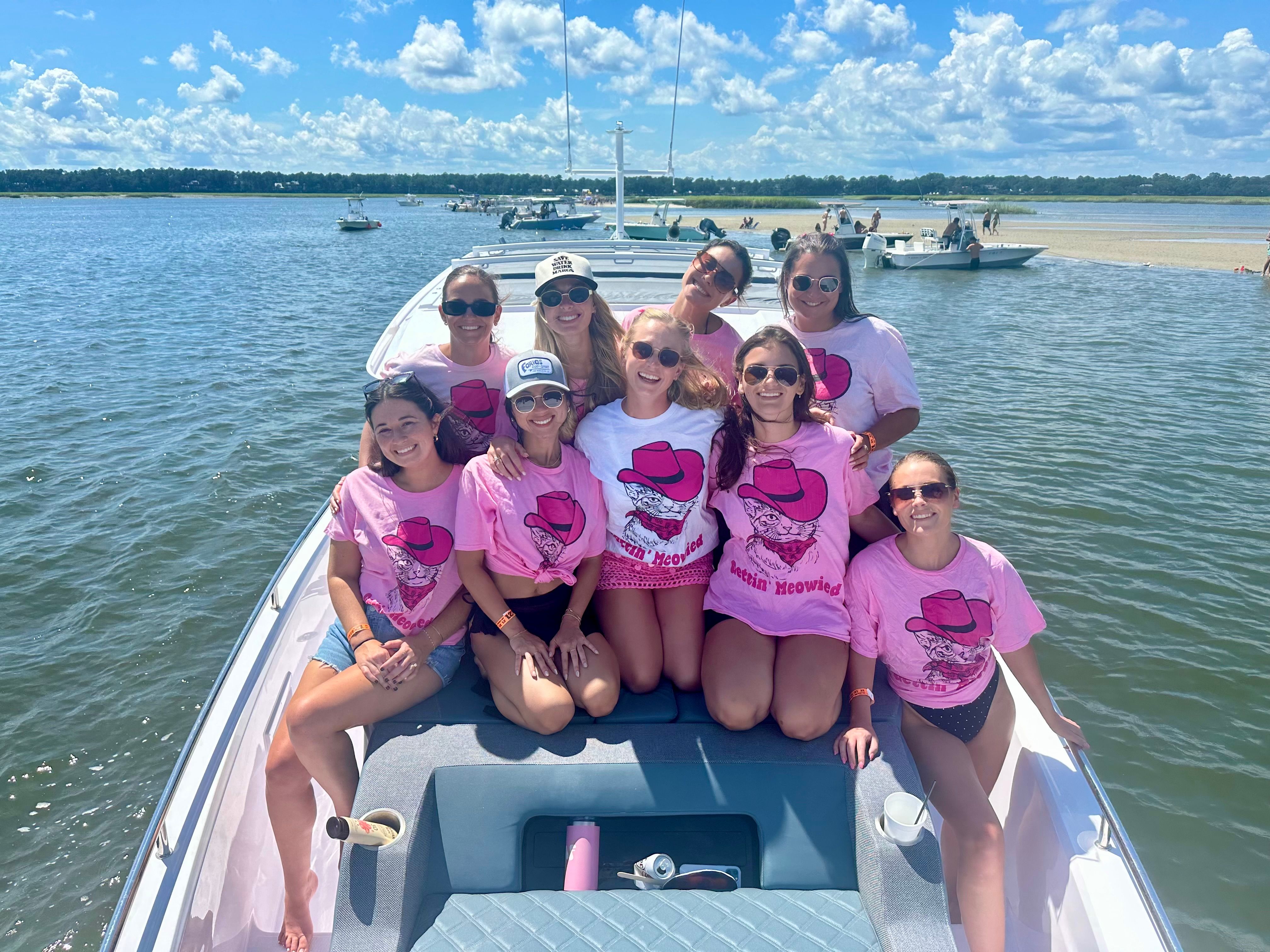 Group of nine friends in matching pink T-shirts gathered on the bow of a white motorboat in a sunny coastal inlet, with nearby anchored boats, a sandbar and blue sky with fluffy clouds in the background.
