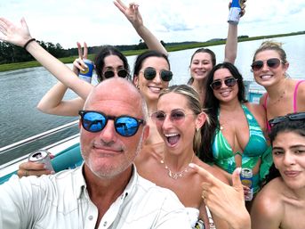 Boat-party group selfie on a coastal marsh river—smiling friends in swimsuits and sunglasses holding canned drinks during a sunny summer outing.