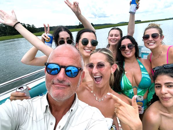 Boat-party group selfie on a coastal marsh river—smiling friends in swimsuits and sunglasses holding canned drinks during a sunny summer outing.