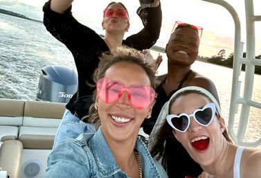 Four friends on a motorboat at sunset wearing heart-shaped sunglasses, smiling and posing for a playful selfie with water and shoreline behind them.